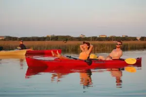 Two kayakers relaxing in a red tandem kayak on calm marsh water at sunset in Murrells Inlet
