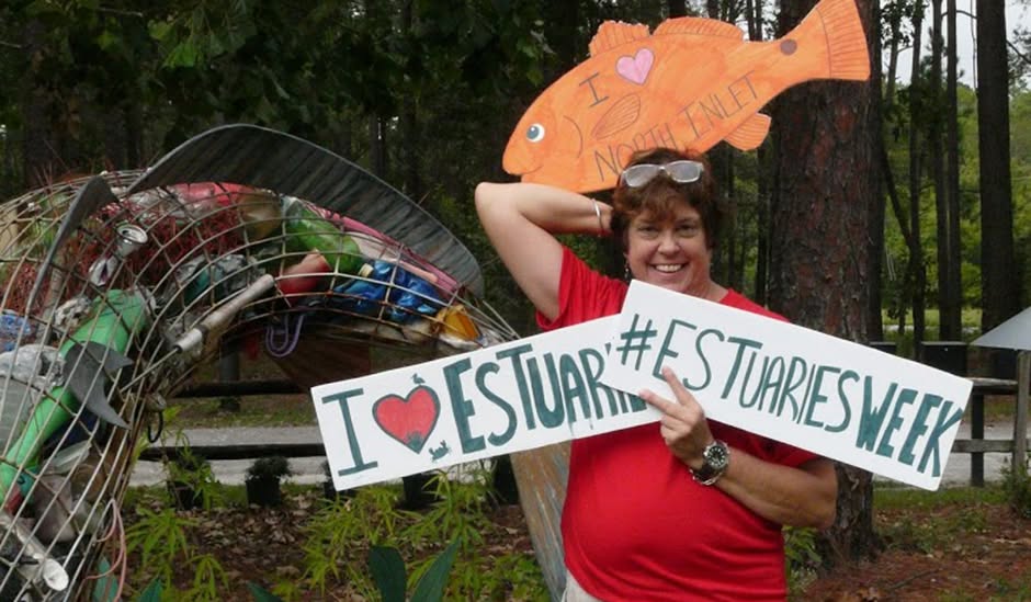 Volunteer holding I Love Estuaries and #EstuariesWeek signs beside a fish sculpture made of collected litter